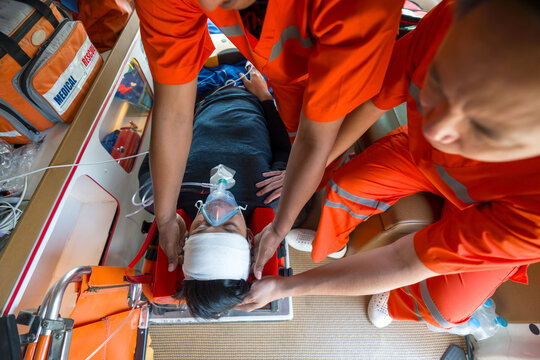 A person is wearing an oxygen mask, getting help from paramedic in orange uniform inside an ambulance. Top view