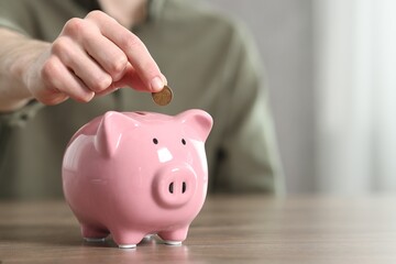 Financial savings. Man putting coin into piggy bank at wooden table, closeup. Space for text