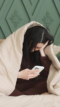 Worried Caucasian woman texting in online social media app, using phone sitting on bed and wrapped in blanket. Spending pastime concept. Vertical video.
