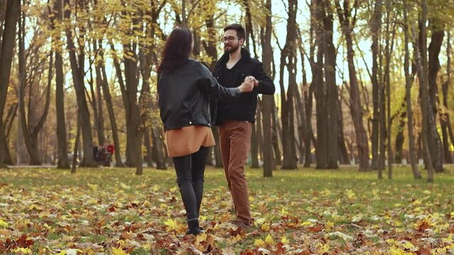 Young loving couple walking outside in the city park in sunny weather, dancing on orange yellow red maple leaves, hugging smiling kissing laughing spending time together. Autumn, fall season 