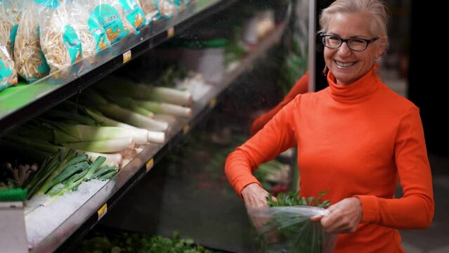 Portrait of smiling happy mature woman shopping for groceries picking green cilantro in grocery store.