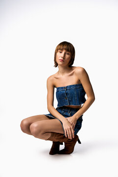 A young woman with short hair is kneeling down in a denim dress in a studio setting.