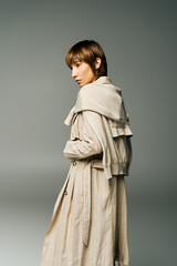 A young woman with short hair stands confidently in a trench coat against a solid gray background.