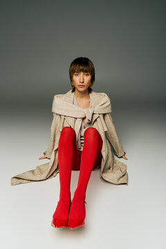 A young woman with short hair elegantly sits on the floor wearing striking red stockings in a studio setting.