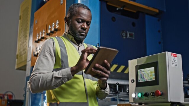 Hardworking multi-ethnic specialists in vests servicing mechanical equipment in large factory. Busy man with his beautiful co-worker checking mechanism while using tablets devices. Industrial concept