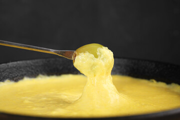 Dipping grape into fondue pot with tasty melted cheese against dark gray background, closeup