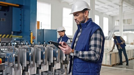 Close-up portrait of focused middle-aged Caucasian factory employee checking tablet information. Purposeful manufacture specialist worker in helmet analyzing production process.