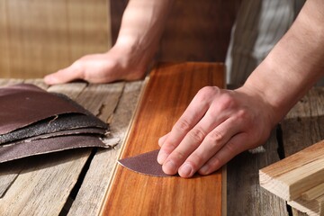 Man polishing wooden plank with sandpaper at table indoors, closeup