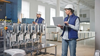 Caucasian female wearing vest and helmet for protection. Standing next to foil production line. Checking data while recording numbers on laptop. Man coming behind with digital device.