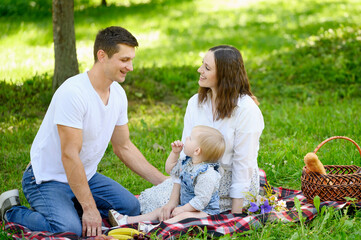 Happy family with toddler having picnic in park in summer. Mom and dad are sitting with child on grass in park, eating healthy food, fruits outdoors, talking. Concept of family vacation, weekend