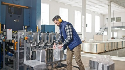 People walking in background. Caucasian male wearing yellow headphones for noise protection. Taking packages of foil. Finishing production line. Working in factory. Busy person. Industry concept.