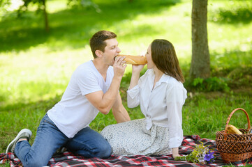  Couple in love on romantic picnic in park in summer, having fun and having bite together, eating French baguette. Concept for vacation, snack while traveling, happy family time. Valentine's Day