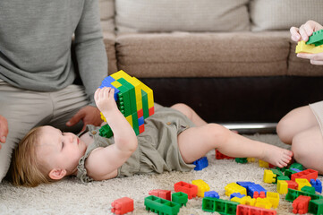 Tired child lies on carpet and holds colorful plastic construction set in his hands,parents sit nearby and play with toddler. Educational entertaining game for children, leisure time Happy childhood.
