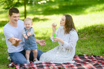 Happy parents and their child are having fun together outdoors, blowing bubbles. Smiling family with toddler enjoying playing with bubbles in park on weekend. Holidays with family.