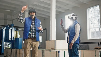 Close up of two male middle-aged engineers in headset and blue vests standing in factory and having wonderful virtual reality experience. Indoors. Men in VR goggles moving their hands in air. - obrazy, fototapety, plakaty