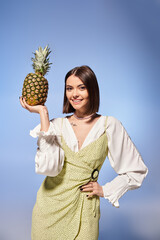 A young woman with brunette hair joyfully holds a pineapple up to her face in a studio setting.