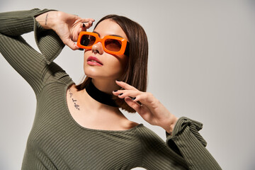 A young woman with brunette hair wearing a green shirt and orange sunglasses poses in a studio...