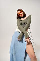 A young woman with brunette hair strikes a stylish pose in a studio while wearing a skirt and...