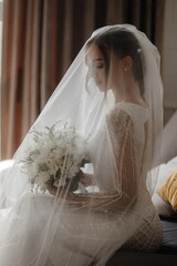 Elegant bride with a white bouquet of flowers sits on a bed and looks away