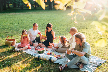 .Big family sitting on the picnic blanket in city park during weekend Sunday sunny day. They are smiling, laughing and eating boiled corn and watermelon. Family values and outdoors activities concept.