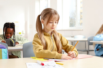 Little schoolgirl writing in the exercise book with classmates on the background in classroom
