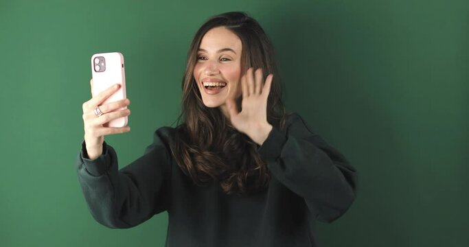 Happy brunette woman having mobile video call, wave hands and say hi, hello isolated on green background. Nice to see you. Happy emotions. Beautiful young girl speaking video call online smartphone.