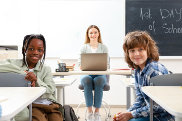 Smiling teacher sitting at the desk, using laptop while kids looking at the camera in classroom