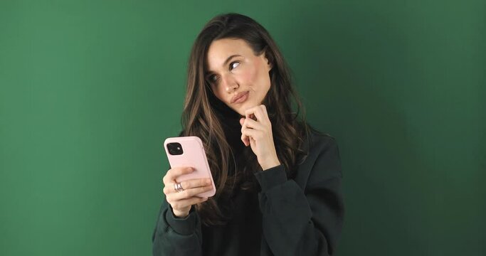 Smiling beautiful young woman isolated on green background. Girl in green sweatshirt flirting while chatting on mobile phone, dreaming. Woman think about answer, bite lip, look at side.