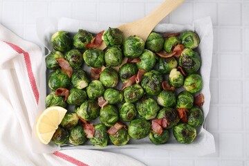 Delicious roasted Brussels sprouts, bacon, lemon and wooden spatula on white tiled table, top view