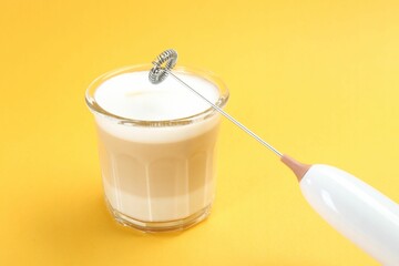 Mini mixer (milk frother) and tasty cappuccino in glass on yellow background