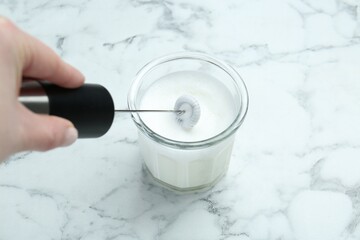 Woman whisking milk in glass with mini mixer (milk frother) at white marble table, closeup © New Africa