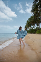Woman spins with a smile on a sandy tropical beach, her blue dress flowing, with palm trees in the background