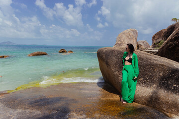 Woman in a green dress stands by a large rock on a tranquil tropical beach, gazing out at the sea