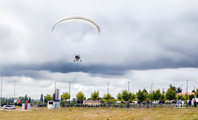 Paramotor with white paraglider flies in the sky over the field on a cloudy day.