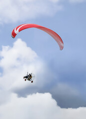 Person on a paramotor with red paraglider flying among the clouds of the blue sky.