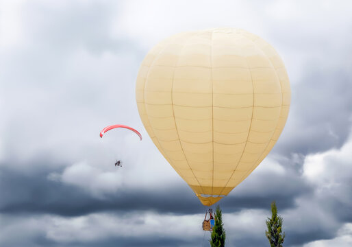 Large hot air balloon flying in the sky with clouds next to a paramotor at an air festival.