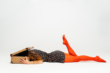 Woman Lies Flat on Floor With Head Buried in Suitcase and Leg Elevated Against White Backdrop