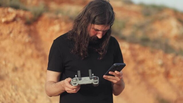 A brutal man with long hair and a beard is learning to fly a drone with a drone controller in his hands and a phone, studying, technology, discoveries, drone flight, test drive, nature, explore