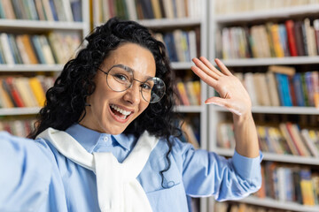 Joyful Hispanic woman with glasses taking a selfie in a university library, surrounded by...