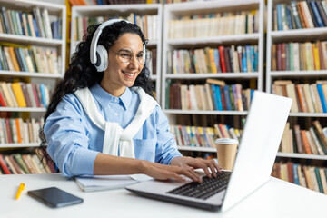 Smiling woman with headphones studying at a laptop in a library setting, surrounded by books and a coffee cup.