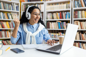 Joyful young student with headphones celebrating academic success in front of a laptop at the...