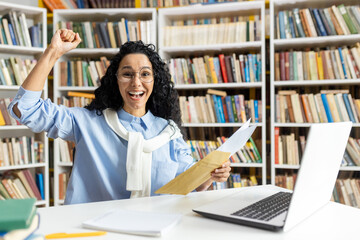 Joyful woman in glasses celebrates as she receives a positive postal message, raising her fist in triumph at a library.