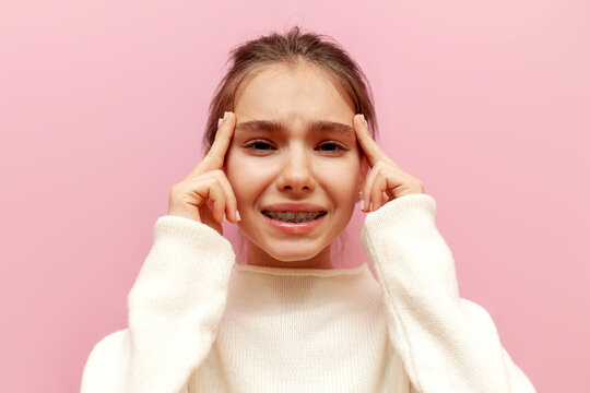 unhappy teenage girl with braces suffers from headache and migraine on pink isolated background, sad child holding hands on head and getting sick