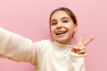 cheerful teenage girl with braces taking selfie and showing peace sign on pink isolated background,...
