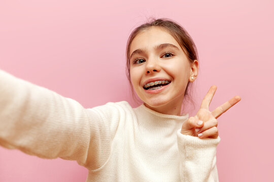 cheerful teenage girl with braces taking selfie and showing peace sign on pink isolated background, happy child taking photo of herself and showing two fingers