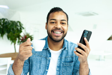 cheerful african american man with braces holds a cup of coffee and a smartphone in a white cafe, a man in a denim shirt drinks coffee in the morning and uses a mobile phone online