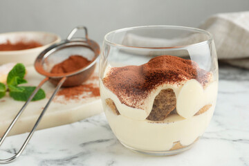 Delicious tiramisu in glasses and sieve with cocoa powder on white marble table, closeup