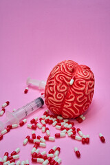 Red human brain with two syringes and a pile of red and white pills and capsules on a pink table.