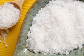 Natural sea salt in bowl and spoon on yellow wooden table, closeup