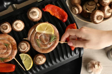  Woman cooking homemade sausages with mushrooms and bell pepper on electric grill at wooden table, top view © New Africa